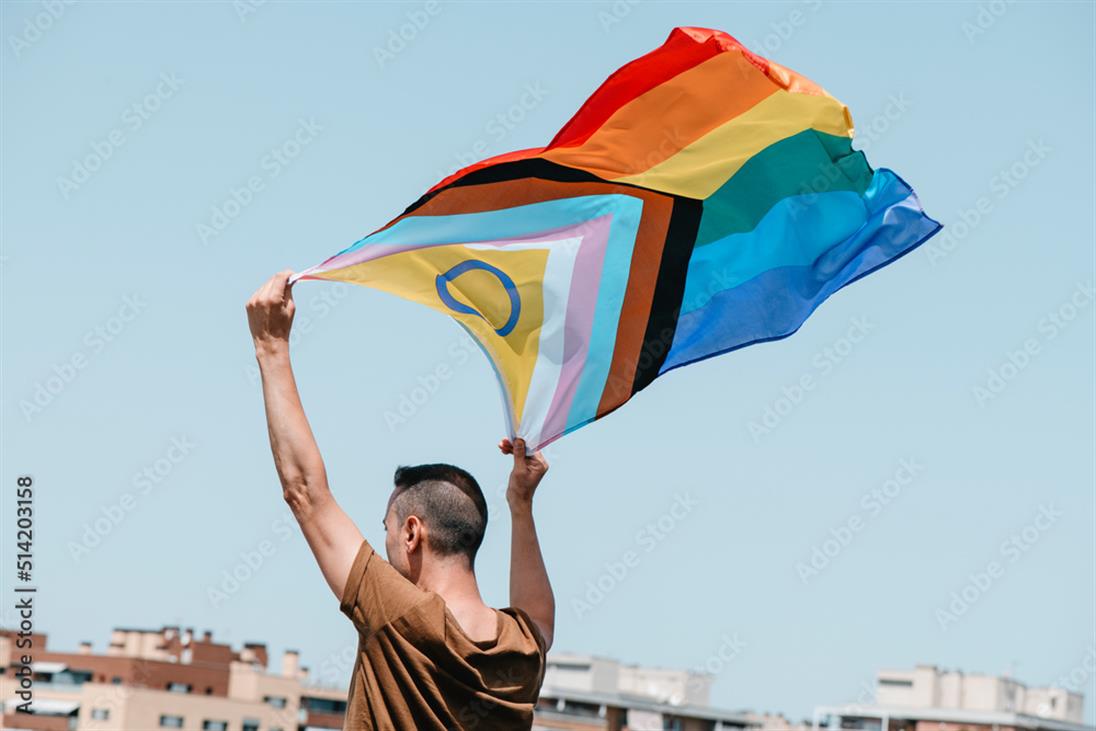 Eine männliche aussehende Person von hinten, welcher eine progressive Regenbogenflagge in den Himmel hebt. Diese weht im Wind, sodass man die gesamte Flagge sieht und im Hintergrund den blauen Himmel.